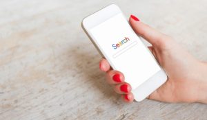 Mão segurando um celular em que na tela aparece a palavra Search em letras coloridas, que remetem as cores do Google.